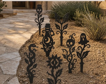 May include: A set of seven black metal garden stakes featuring silhouettes of a skull, a potion bottle with a moon, a bat, a snake, an eye-centred flower, a skull on a vine, and a ghost. The text reads SOLD INDIVIDUALLY OR AS A SET OF 7.