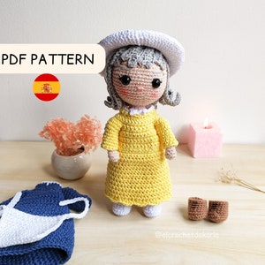 Puede incluir: Patrón de muñeca de crochet en PDF en español. La muñeca lleva un vestido amarillo y un sombrero blanco. La muñeca está sentada sobre una superficie de madera con un pequeño ramo de flores y una vela en el fondo.