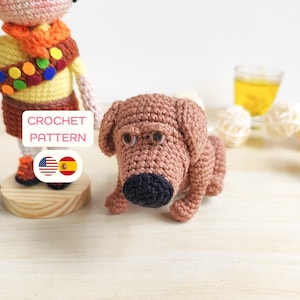 Puede incluir: Un perro marrón tejido a ganchillo con una nariz negra y ojos marrones. El perro está acostado. La imagen también incluye una figura tejida a ganchillo de una persona y el texto "CROCHET PATTERN".