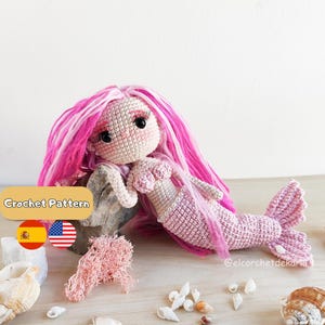May include: A crocheted mermaid doll with pink and white hair, a pink tail, and a seashell top. The doll is lying on a rock with a few seashells and coral around it. The text "Crochet Pattern" is visible.