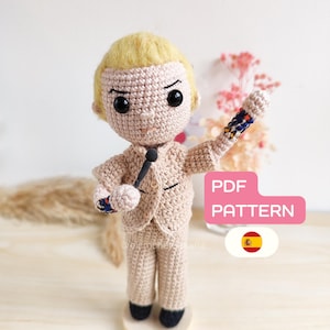 Puede incluir: Muñeco de ganchillo de un hombre con traje beige, sosteniendo un micrófono negro y con pelo rubio. El muñeco tiene ojos negros y está de pie. El texto "PDF PATTERN" está en un rectángulo rosa. También se ve una bandera española.