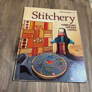 May include: A vintage "Stitchery" book with a cover featuring embroidery examples, a doll, and a floral embroidery hoop. The book's title is in a large, red font. The cover also includes the words "Embroidery, Applique, Crewel."