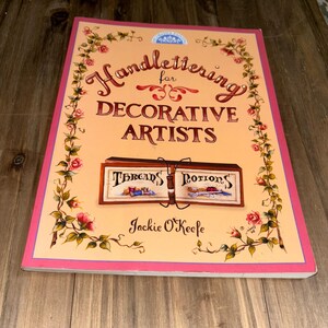May include: A book titled "Handlettering for Decorative Artists" by Jackie O'Keefe. The cover features a pink and yellow background with a floral border. The title is written in a decorative script font. The book is about hand lettering for decorative artists.