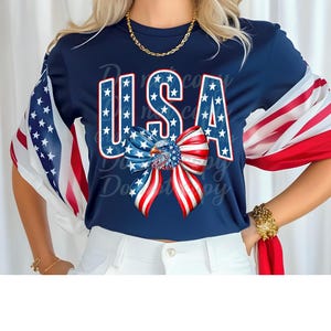 May include: Navy blue t-shirt with the word "USA" in white outlined letters filled with blue stars. A red, white, and blue bow is below the text. The sleeves feature a flowing American flag design.