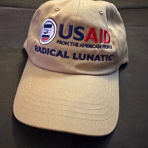 May include: Tan baseball cap with the text "USAID FROM THE AMERICAN PEOPLE" in red and blue embroidery. The cap also features the words "RADICAL LUNATIC" in blue. A circular logo is on the left side of the cap.