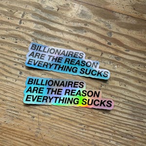 Könnte beinhalten: Zwei holografische Aufkleber mit dem Text "BILLIONAIRES ARE THE REASON EVERYTHING SUCKS" in fetter, serifenloser Schrift. Die Aufkleber haben eine rechteckige Form mit abgerundeten Ecken und liegen auf einer Holzoberfläche.