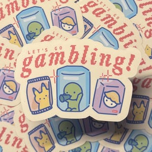 Puede incluir: Una pila de pegatinas con un dise&ntilde;o de dibujos animados. Las pegatinas presentan un fondo amarillo claro con un texto pixelado rojo que dice "Let's go gambling!" Las pegatinas tienen un dise&ntilde;o de dibujos animados de una criatura verde, una criatura amarilla y una persona rubia en una caja.