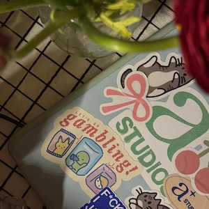 Puede incluir: Un port&aacute;til azul con varias pegatinas. Las pegatinas incluyen un gato con un lazo, una cinta rosa, un logotipo verde y blanco que dice "Studio", una pegatina que dice "Let's go gambling!" con una imagen de dibujos animados de un pato en un frasco de vidrio, una pegatina azul que dice "F*ck you, I just try", y una pegatina que dice "a+ studios".