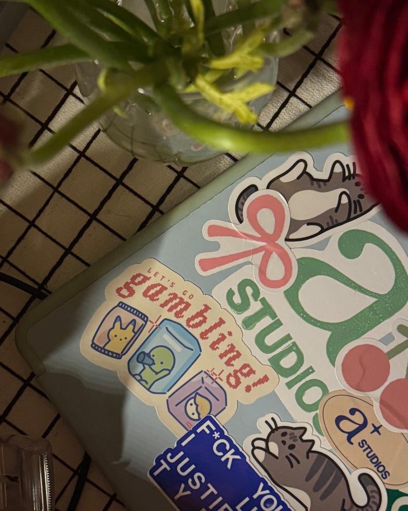 Puede incluir: Un port&aacute;til azul con varias pegatinas. Las pegatinas incluyen un gato con un lazo, una cinta rosa, un logotipo verde y blanco que dice "Studio", una pegatina que dice "Let's go gambling!" con una imagen de dibujos animados de un pato en un frasco de vidrio, una pegatina azul que dice "F*ck you, I just try", y una pegatina que dice "a+ studios".