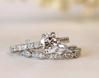 May include: A set of two silver engagement rings with a large round center stone and smaller stones set in a pave setting. The band of the larger ring is a simple band with stones set in a pave setting. The band of the smaller ring is a delicate band with stones set in a pave setting.