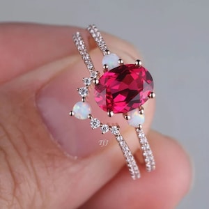 May include: A rose gold ring set featuring a large oval-cut ruby center stone. The set includes a curved band with small white opal accents and a second band with small clear stones.