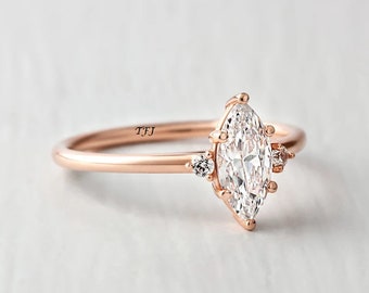 Pode incluir: Um anel de noivado em ouro rosa com uma pedra central em forma de marquise e duas pedras de acento menores. O anel é gravado com as iniciais "TFJ".