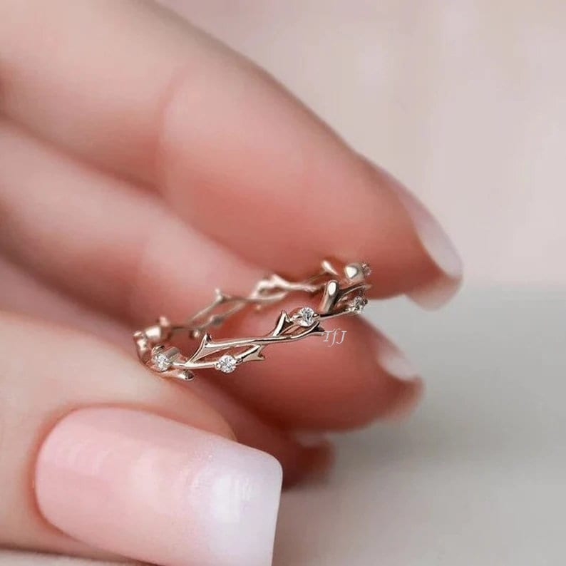 May include: A delicate gold ring with a twig design and small clear stones. The ring is on a finger with pink nail polish.