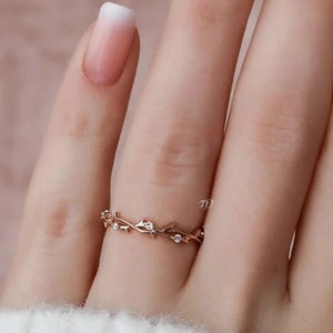 May include: A delicate gold ring with a vine design and small clear stones. The ring is on a finger with a white and pink ombre nail polish.