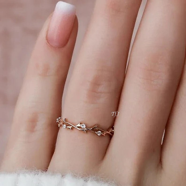 May include: A delicate gold ring with a vine design and small clear stones. The ring is on a finger with a white and pink ombre nail polish.
