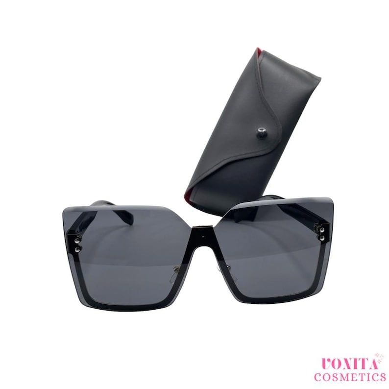 Op de afbeelding: Zwarte oversized zonnebril met een bijpassende zwarte etui. De zonnebril heeft een vierkant montuur en donkere glazen. De etui is van zacht, zwart leer met een rode binnenkant. De merknaam "ROXITA COSMETICS" is roze gedrukt.