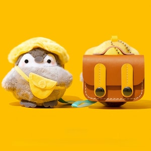 May include: A soft penguin toy with a yellow hat and satchel, alongside a brown leather bag with yellow details. The background is a bright yellow.