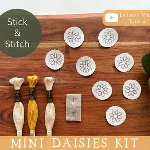 May include: A craft kit with the text "Stick & Stitch" and "Mini Daisies Kit." The kit includes daisy-shaped fabric patches, embroidery floss in cream and yellow, and a small felt piece. A video tutorial is included.