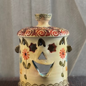 May include: A ceramic candle holder with a whimsical face, featuring a smiling mouth, star-shaped eyes, and floral details. The holder is painted in shades of brown, red, green, and yellow.