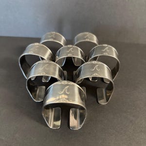 May include: A collection of eight silver napkin rings. Each ring is engraved with the letter "A" in a decorative script. The rings have a curved design with a slot on one side, and are arranged on a dark surface.