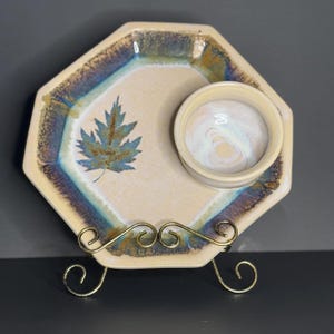 May include: A ceramic serving dish with a small dipping bowl. The dish is octagonal and has a light beige background with a blue and green leaf design. The dipping bowl is white with a light beige interior. The dish is on a gold metal stand.