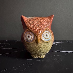 May include: A ceramic owl figurine featuring a textured, scalloped feather pattern. The upper portion is a warm orange-brown, while the lower body is a lighter, muted yellow-beige. It has two large, circular eyes with white centers.