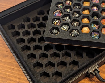 May include: A black metal carrying case featuring a removable foam insert with a honeycomb grid pattern. The case contains various multi-sided gaming dice in assorted colors, including orange, black, and glittered finishes, stored in individual slots.