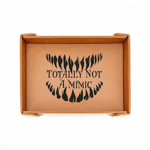 May include: A rectangular brown tray with the text "TOTALLY NOT A MIMIC" in black. The tray has a stitched border and small corner tabs. The text is above a stylized toothy grin.