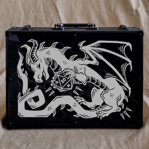 May include: Black case with a silver dragon graphic holding a 20-sided die. The dragon has large wings and is surrounded by flames. The case has a handle and metal accents.