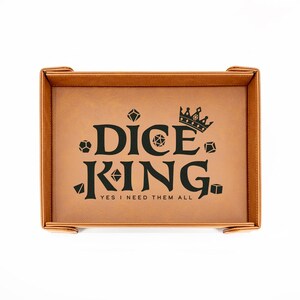 May include: A rectangular, tan dice tray with a black graphic that reads "DICE KING" with a crown and dice illustrations. The text "YES I NEED THEM ALL" is below the main text. The tray has raised edges and corner tabs.