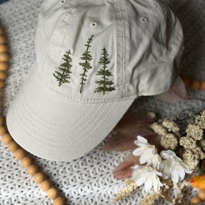May include: A beige baseball cap featuring an embroidered design of three green pine trees on the front panel. The cap has a curved brim with stitched detailing and a button at the top center, constructed from a solid light-colored fabric.