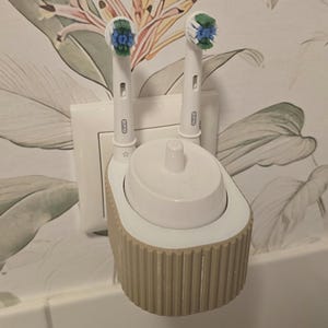 May include: A white and beige plastic electric toothbrush charging stand with a ribbed exterior design. The stand holds two Oral-B toothbrush heads with blue and green bristles, and features a central charging port for a toothbrush handle.