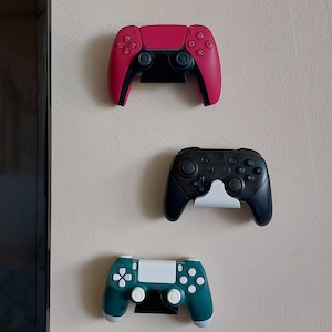 Könnte beinhalten: Drei Videospielcontroller, die an einer Wand montiert sind. Die Controller sind in den Farben Pink, Schwarz und Teal-Grün.