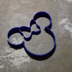 Puede incluir: Un cortador de galletas de plástico azul con forma de la cabeza de Mickey Mouse. El cortador está sobre una superficie marrón claro con harina blanca esparcida alrededor.