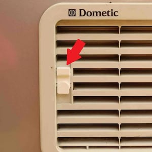 Könnte beinhalten: Nahaufnahme einer beigefarbenen Dometic-Lüftungsabdeckung mit horizontalen Lamellen. Ein weißer rechteckiger Riegel ist auf der linken Seite sichtbar, und das Dometic-Logo befindet sich in der oberen linken Ecke. Der Hintergrund ist eine hellbeige Oberfläche mit blauen Streifen.