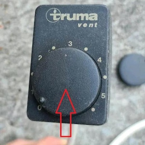 May include: A black Truma vent control panel with a circular dial and numbers 0-5. The Truma logo is visible at the top. A separate black cap is to the right.