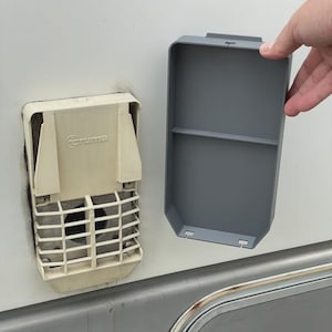 Könnte beinhalten: Nahaufnahme einer Truma-Wohnmobil-Ofenlüftung und einer grauen Zugangsklappe. Die Lüftung ist cremefarben mit einem Gitter und dem Truma-Logo. Die graue Platte ist rechteckig mit einem Regal.