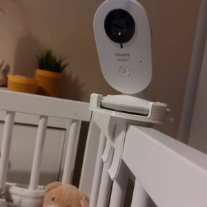 May include: White baby monitor with a black camera lens, mounted on a white cot rail. The monitor has the text "PHILIPS AVENT" on it. The text "Variante 1" is visible in the lower right corner of the image.