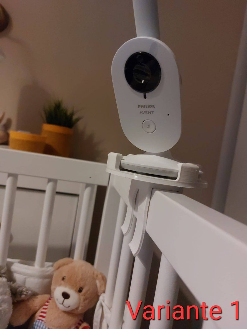 May include: White baby monitor with a black camera lens, mounted on a white cot rail. The monitor has the text "PHILIPS AVENT" on it. The text "Variante 1" is visible in the lower right corner of the image.