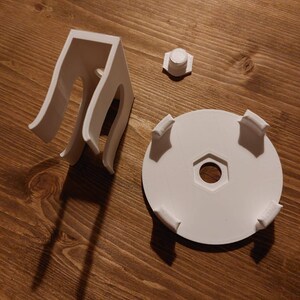 May include: White 3D printed parts for a project, including a base with four prongs, a small round piece, and a tall, open-ended structure with two loops.