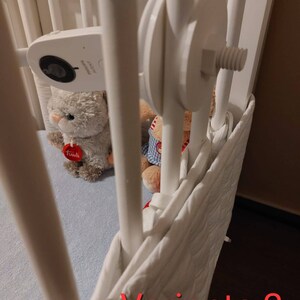 May include: A white cot with a white cot bumper and a white baby monitor attached to the side. There are two stuffed toys in the cot, one grey and one red. The text "Variante 2" is written in red at the bottom of the image.