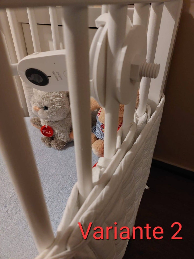 May include: A white cot with a white cot bumper and a white baby monitor attached to the side. There are two stuffed toys in the cot, one grey and one red. The text "Variante 2" is written in red at the bottom of the image.
