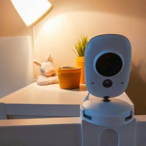 Könnte beinhalten: Ein weißer Babymonitor mit einem schwarzen Objektiv ist an einem weißen Babybettgitter befestigt. Der Monitor ist nach vorne gerichtet und das Objektiv zeigt auf das Babybett. Der Monitor ist über ein weißes Kabel mit einer Stromquelle verbunden. Das Babybett steht in einem Raum mit einer Lampe auf einem Tisch im Hintergrund. Der Text "Variante 1" ist in der linken unteren Ecke des Bildes sichtbar.