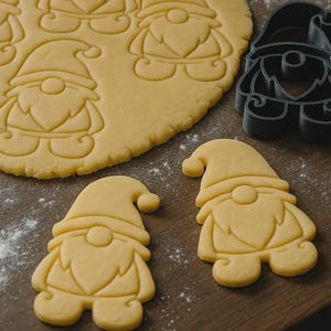 Puede incluir: Primer plano de galletas con forma de gnomo y un cortador de galletas. Las galletas están cortadas de masa, con un cortador de plástico negro cerca. La escena está ambientada sobre una superficie de madera, con harina espolvoreada.