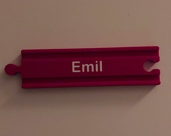 May include: A pink wooden train track piece with the name "Emil" printed in white on the top.