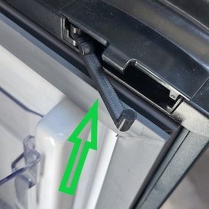 Könnte beinhalten: Nahaufnahme eines schwarzen Kühlschrank-Türverschlusses mit einem weißen Pfeil, der auf den Verschluss zeigt. Der Verschluss ist aus Kunststoff und hat einen kleinen Metallstift.