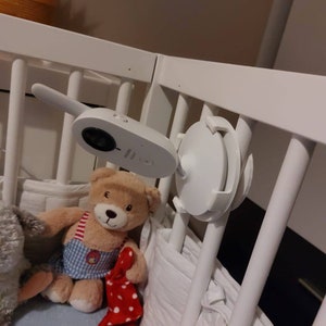 May include: A white baby monitor with a camera mounted on a white cot. The monitor is attached to the cot with a white plastic mount. A teddy bear with a red and white polka dot blanket is in the cot.