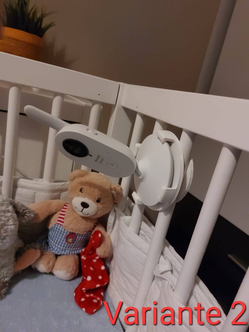 May include: A white baby monitor with a camera mounted on a white cot. The monitor is attached to the cot with a white plastic mount. A teddy bear with a red and white polka dot blanket is in the cot.