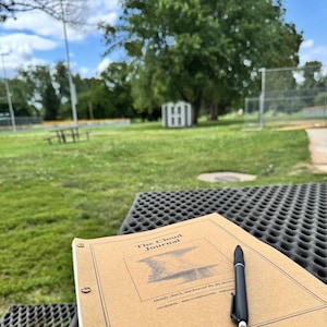 May include: A tan-coloured journal titled The Cloud Journal with a black pen resting on its cover. The cover features a central rectangular illustration of a cloud formation and text that reads Identify, sketch, and forecast the sky above.