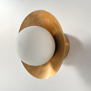 May include: A modern wall sconce featuring a spherical white glass globe set within a curved, brass-toned metal shade. The fixture is mounted on a white wall, showcasing a minimalist design.